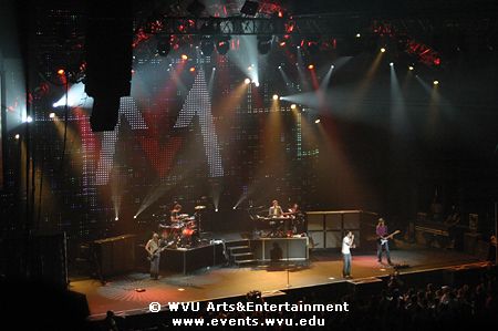 Wide shot of Maroon 5 on stage with spotlights casting grey and red halos.