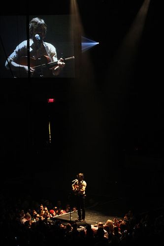 Wide shot of stage with Scott Avett in the foreground and a close up shot of him on the video screen in the upper left corner. 