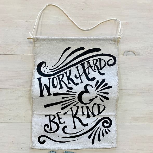 Hanging banner made from white canvas with the words "work hard, be kind" painted on it in black.