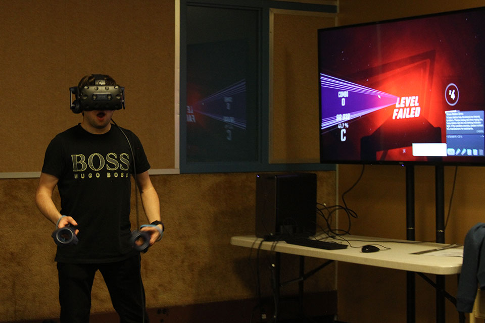 A student stands next to a large screen TV wearing a virtual reality gaming headset and holding gaming controllers. Photo by Hannah Wicks.