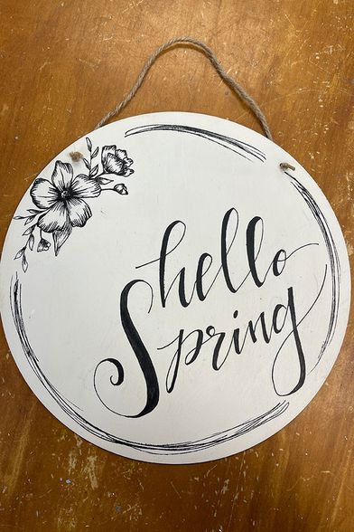white, round, wooden door hanger with "hello spring" painted on it in black script. Twine is affixed to it for hanging.