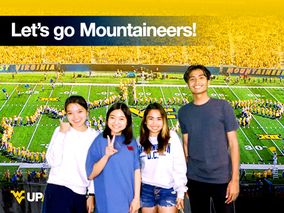 A group of four students pose for a photo with the Pride of West Virginia forming the State of West Virginia on the football field in the background.
