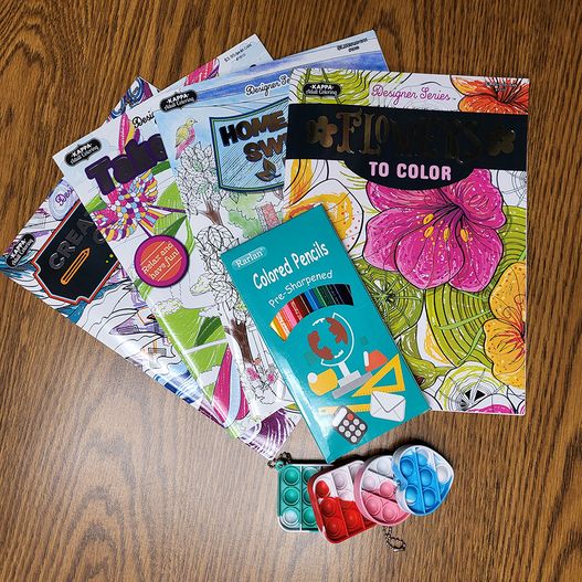 various adult coloring books fanned out on a table with a box of colored pencils and four fidget pop-it keychains in various shapes and colors.