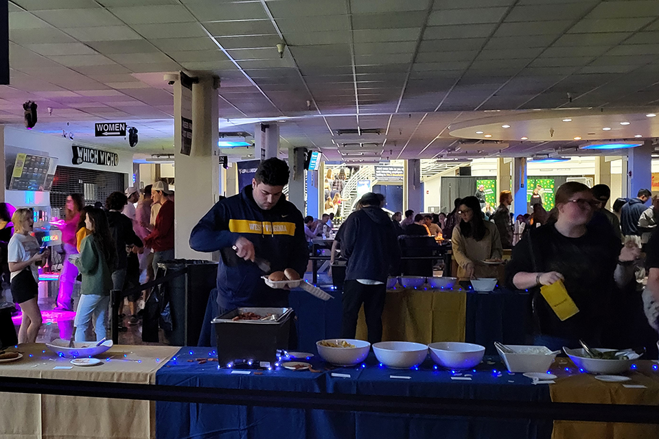 A male student wearing a West Virginia University sweatshirt fills his food container while other students wait their turn in the buffet line.