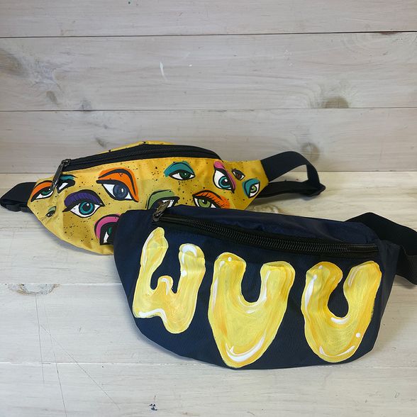 Two painted fanny packs. One is blue with the letters W, V and U painted in yellow. The other is yellow with eyes painted on it.