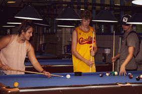 A female student lines up her pool cue while two male students look on.