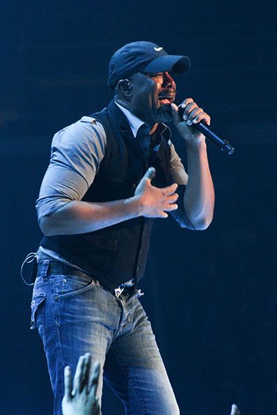 Darius Rucker sings on stage in 2014 wearing jeans, a dark colored vest and a black baseball hat
