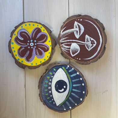Round slices of wood with illustrations painted on them. One is yellow with a purple flower. One is brown with white mushrooms and one depicts an eye in blue and white.