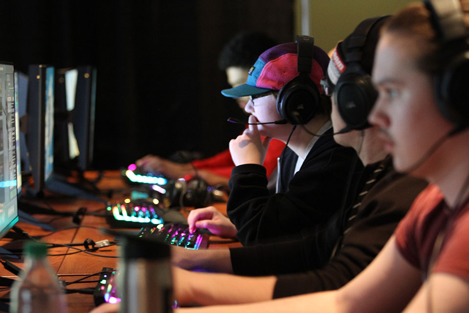 Students wear headsets while sitting in front of computer monitors playing computer games.